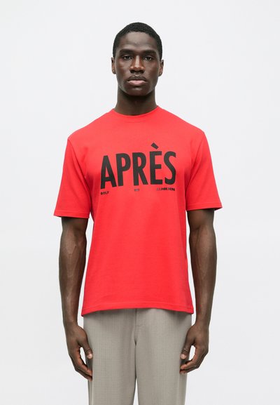 Homme portant un T-shirt rouge avec le texte noir "APRÈS" et un pantalon gris, se tenant devant un fond blanc uni.