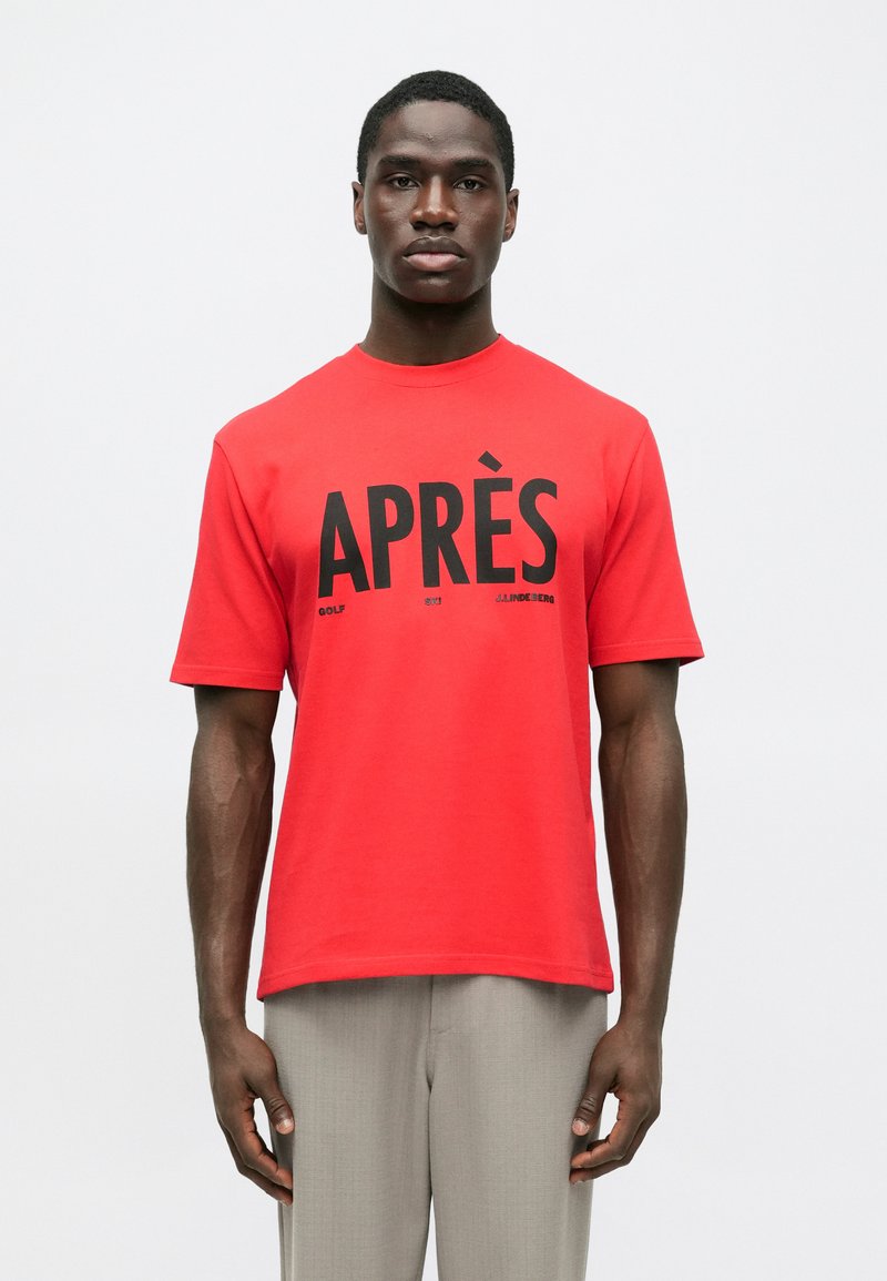 Άνδρας με κόκκινο T-shirt και μαύρο κείμενο "APRÈS" και γκρίζους παντελόνες, στέκεται μπροστά σε λευκό απλό φόντο.