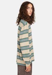 Camicia beige a maniche lunghe con strisce orizzontali verdi e blu. Vestibilità comoda con scollatura a girocollo e polsini affusolati. Indossata con pantaloni grigi.