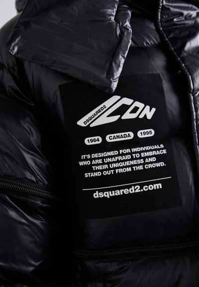 Veste noire matelassée avec une finition brillante, fermeture éclair, et une étiquette avec texte et graphiques blancs, fournissant des informations sur la marque et le design.