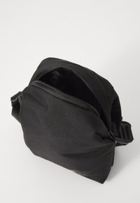 Kompakte schwarze Tasche aus strukturiertem Stoff mit einem Reißverschluss. Sie verfügt über einen verstellbaren Gurt und ein kleines Logo an der Seite.