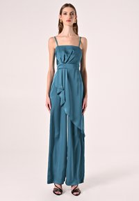 Tutina in satin teal con spalline sottili, dettaglio drappeggiato sul davanti e gambe ampie. Abbinata a sandali neri con tacco, che mostrano una texture liscia e lucida.