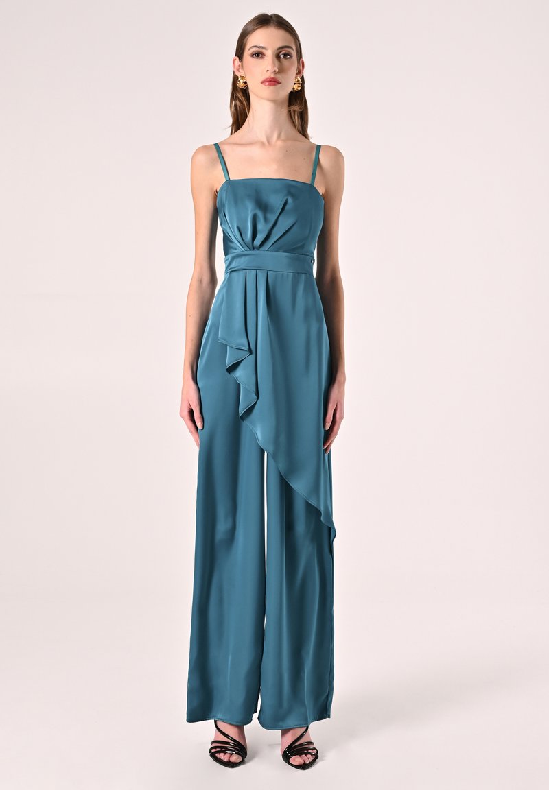 Tutina in satin teal con spalline sottili, dettaglio drappeggiato sul davanti e gambe ampie. Abbinata a sandali neri con tacco, che mostrano una texture liscia e lucida.
