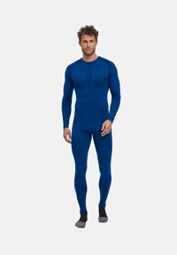 Haut de sport à manches longues bleu et leggings assortis, fabriqués en tissu extensible avec des accents texturés. Le modèle se tient sur un arrière-plan uni.