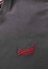 T-shirt in cotone grigio scuro con scollatura rotonda e un prominente logo ricamato rosso "SuperDry" sul davanti.