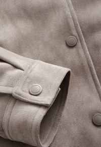 Lichtgrijze suede jas met een gladde textuur, twee grote knopen en een opgerolde manchette met een stiksel langs de rand.