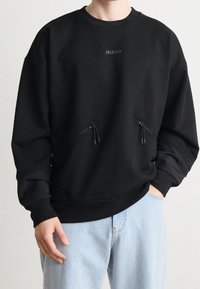 Persona che indossa una felpa nera HUGO con due tasche con cerniera sul davanti e jeans azzurri, mano in tasca.