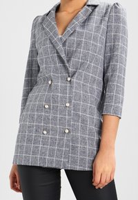 Dubbelknäppt kavaj i grå och vit rutmönster, med pärlknappar och trekvartslånga ärmar, tillverkad av en strukturerad tyg.