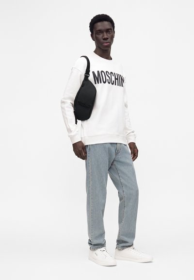Homme portant un sweat blanc Moschino, un jean bleu clair, des baskets blanches et un sac bandoulière noir, debout devant un fond clair uni.