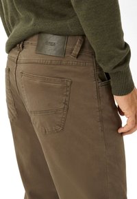 Man in een olijfgroene trui en een paarse broek met achterzakken en een leren merklabel op de tailleband.