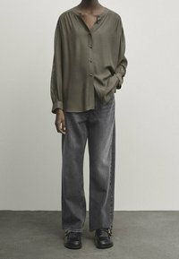 Olive green, button-up shirt with pleated shoulder details, paired with loose-fitting, dark gray jeans and black leather shoes.