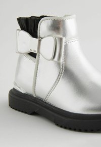 Silberne metallische Knöchelstiefel mit schwarzem Rüschensaum, versehen mit einer Schleifendetail und einer strukturierten Gummisohle mit Grip. Markenname sichtbar.
