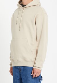 Personne portant un sweat à capuche beige avec une poche avant et un jean bleu, debout contre un fond blanc.