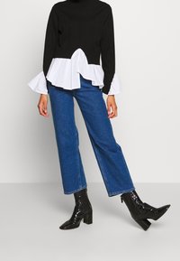 Personne portant un jean bleu droit, des bottines noires à talons et un pull noir avec des poignets et un ourlet à volants blancs.
