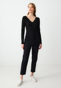 Schwarzes Langarmoberteil mit einem gewellten V-Ausschnitt, kombiniert mit schwarzen geraden Hosen und weißen Turnschuhen. Glatte Textur, minimalistisches Design.