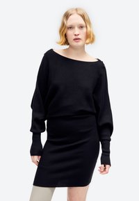 Kobieta o krótkich blond włosach ubrana w czarną prążkowaną sukienkę sweterkową z długim rękawem, stojąca na jednolitym białym tle.