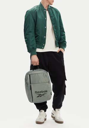 Osoba w zielonej kurtce bomberce, białej koszuli, czarnych spodniach typu cargo, białych trampkach trzymająca po szary plecak Reebok przy boku.