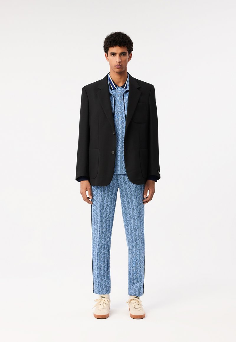 Blazer noir sur une chemise à motif géométrique bleu clair et un pantalon assorti. Baskets crème avec des accents beiges. Tissu lisse, coupe ajustée.