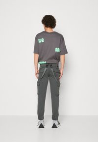 Camiseta gris con texto verde menta, combinada con pantalones cargo grises con correas y zapatillas deportivas grises, vista desde la parte posterior.