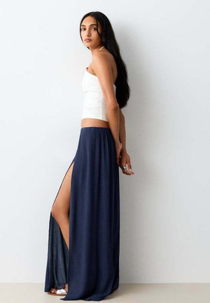 Mujer joven de perfil, vistiendo un top blanco sin tirantes y una falda larga azul marino con una abertura alta, sobre un fondo claro y liso.