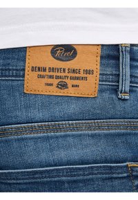 Jeans i denim med en brun läderetikett som har texten "Petrel" och "DENIM DRIVEN SINCE 1989." Blått tyg med gula sömdetaljer.