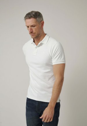 Hombre con una camiseta polo blanca de manga corta y vaqueros oscuros, mirando hacia abajo sobre un fondo claro y liso.