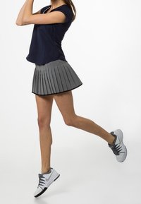 Person klädd i en marinblå topp, grå plisserad kjol och vita sneakers i en mitt-i-steget pose mot en vit bakgrund.