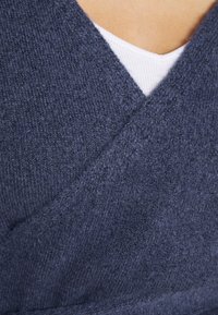 Marinblå stickad sjal med texturerad finish och V-halsdesign. Materialet verkar mjukt med en känsla av komfort, lager på lager över en vit topp.