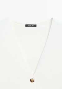 Vêtement blanc à col en V avec un seul bouton marron et une étiquette noire indiquant "Massimo Dutti Fabriqué en Chine" au niveau de l'encolure.