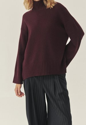 Kvinde iført en mørk burgunder rullekravesweater og sorte, bredbenede bukser med nålestriber, stående mod en ensfarvet baggrund.