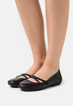 Jalkoja, joissa on mustat nahka-balletit, joissa on ristikkäiset hihnat ja pyöreät napit, seisoo valkoisella pinnalla.