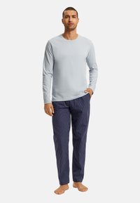 Lichtblauw langemouwen shirt gecombineerd met marineblauwe gestreepte pyjamabroek. De broek heeft een relaxte pasvorm en heeft verticale witte strepen. Blote voeten.