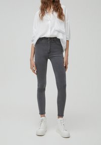 Femme vêtue d'une chemise blanche ample à boutons, d'un jean skinny gris taille haute et de baskets blanches montantes, debout sur un fond uni.