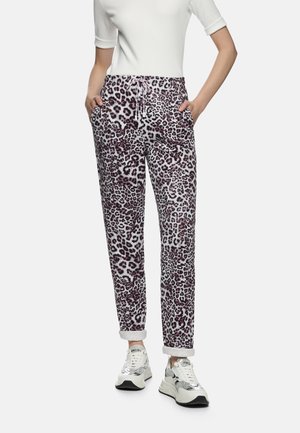 Leopardenmuster-Hose mit weißem Grund, versehen mit lila und schwarzen Mustern, einem Kordelzug in der Taille und aufgekrempelten Bündchen. Kombiniert mit weißen Turnschuhen.