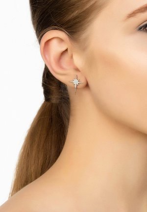 Silver star-shaped earrings with central clear gemstone, featuring pointed accents and a polished surface, worn on an ear.