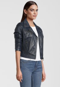 Veste en cuir biker bleu marine avec poches zippées, épaules texturées et col classique. Portée sur une chemise blanche et un jean bleu.