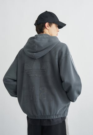 Jeune homme portant un sweat à capuche gris foncé avec un logo Adidas cousu et le numéro 72 dans le dos, une casquette noire et un pantalon noir avec des bandes blanches.