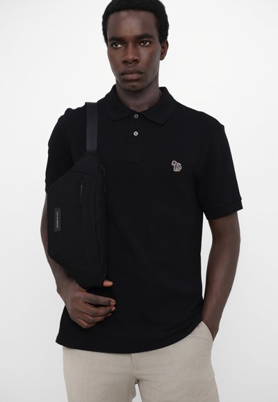 Mustvalge polo särk kolme nööbiga ja brodeeritud logoga, koos heleda beežide pükstega ning musta tõmblukuga rihmakotiga.