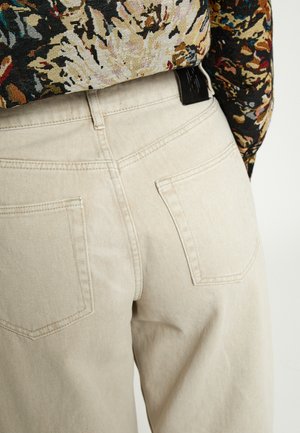 Beežid teksapüksid tagataskutega, mida kannetakse sisse toppitud lillemustrilise pikkade varrukatega särgiga, nähtav vööst allapoole.