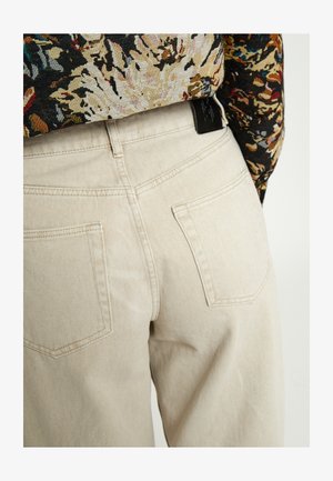 Beige spijkerbroek met achterzakken, gedragen met een ingestopt langermige top met bloemenpatroon, vanaf de taille naar beneden zichtbaar.