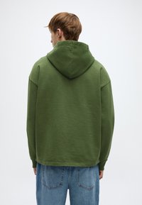 Πράσινο φούτερ με άνετη κουκούλα, χαμηλωμένους ώμους και υφή με ανάγλυφο φινίρισμα. Συνδυάζεται με φαρδιά μπλε τζιν παντελόνια.