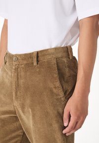 Pantaloni in velluto a coste marroni con righe verticali, dotati di chiusura a bottone, tasche laterali e una vestibilità sartoriale. Indossati con una maglietta bianca.