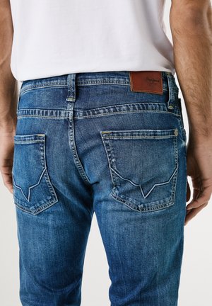 Homme portant un jean en denim bleu avec des poches arrière ornées d'une couture unique et un patch de marque en cuir marron, associé à une chemise blanche.
