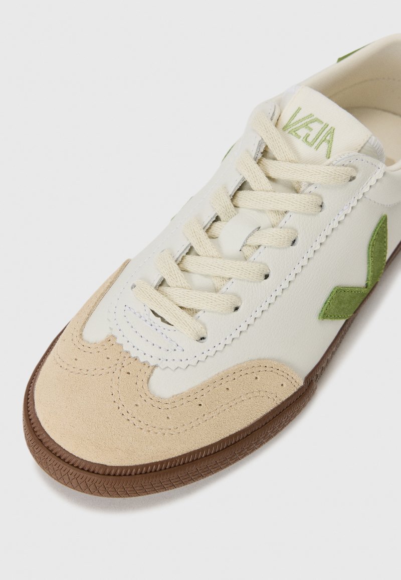 Baskets en cuir blanc avec un bout en suède beige et un logo vert. Équipées de lacets plats crème et d'une semelle en caoutchouc brun avec une adhérence texturée.