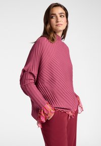 Mujer con cabello largo y castaño, vistiendo un suéter de cuello alto rosa acanalado con puños de volantes estampados y pantalones rojos a juego, con las manos en los bolsillos.