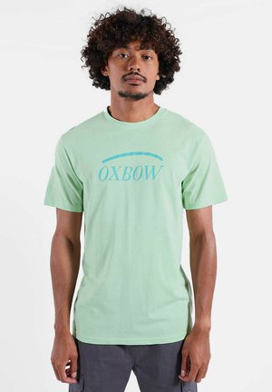 T-shirt en coton vert clair avec un design graphique bleu portant le texte « OXBOW » sur le devant. Manches courtes et coupe classique.