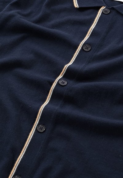 Dunkelblaue Hemdjacke mit weicher Textur, ausgestattet mit einem kontrastierenden hellbeigen Streifen entlang der Knopfleiste und schwarzen Knöpfen.