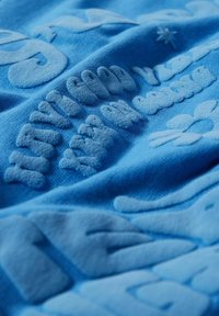 Tissu bleu avec des lettres en relief texturées et des formes décoratives en gros plan montrant des détails de broderie douce et rembourrée.