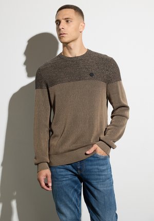 Suéter de punto marrón con una sección superior texturizada, puños acanalados y un sutil logotipo en el pecho, combinado con jeans azules.