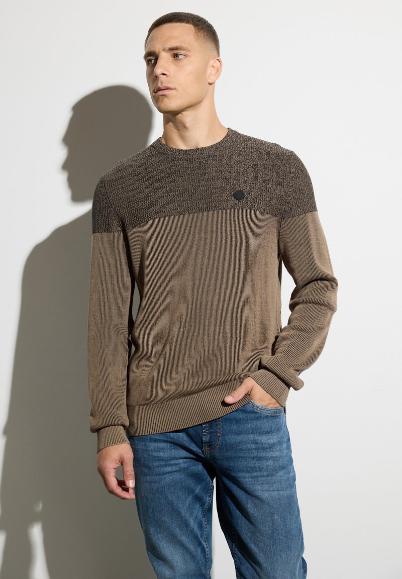 Brauner Strickpullover mit einer strukturierten oberen Sektion, gerippten Bündchen und einem dezenten Logoakzent auf der Brust, kombiniert mit blauen Jeans.
